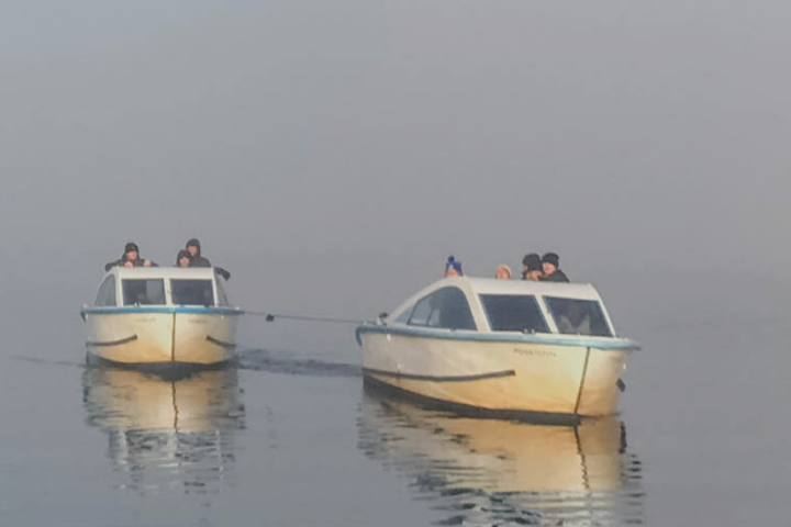 Two small boats with people, connected by a rope, on a misty water surface.