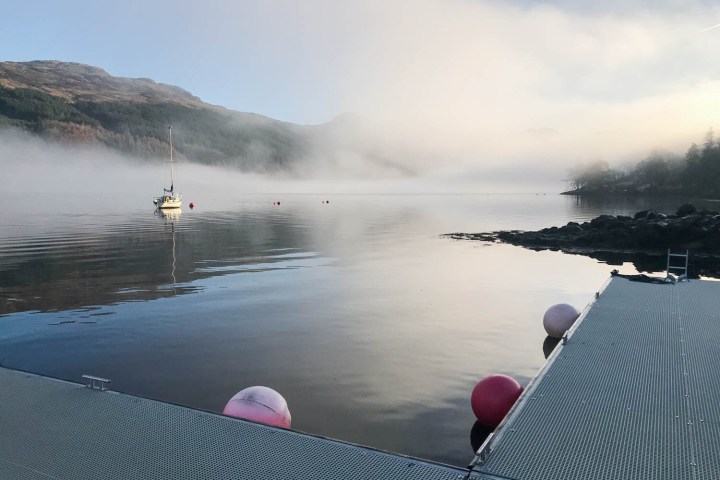 Foggy lake with a dock, buoy, and sailboat next to mountains.