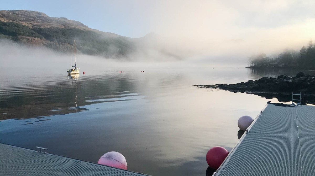 Foggy lake with a dock, buoy, and sailboat next to mountains.