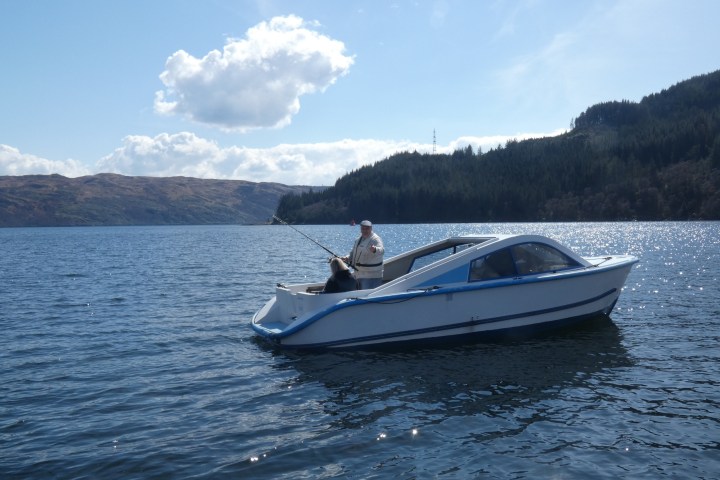Person fishing on a small boat in a lake surrounded by hills.