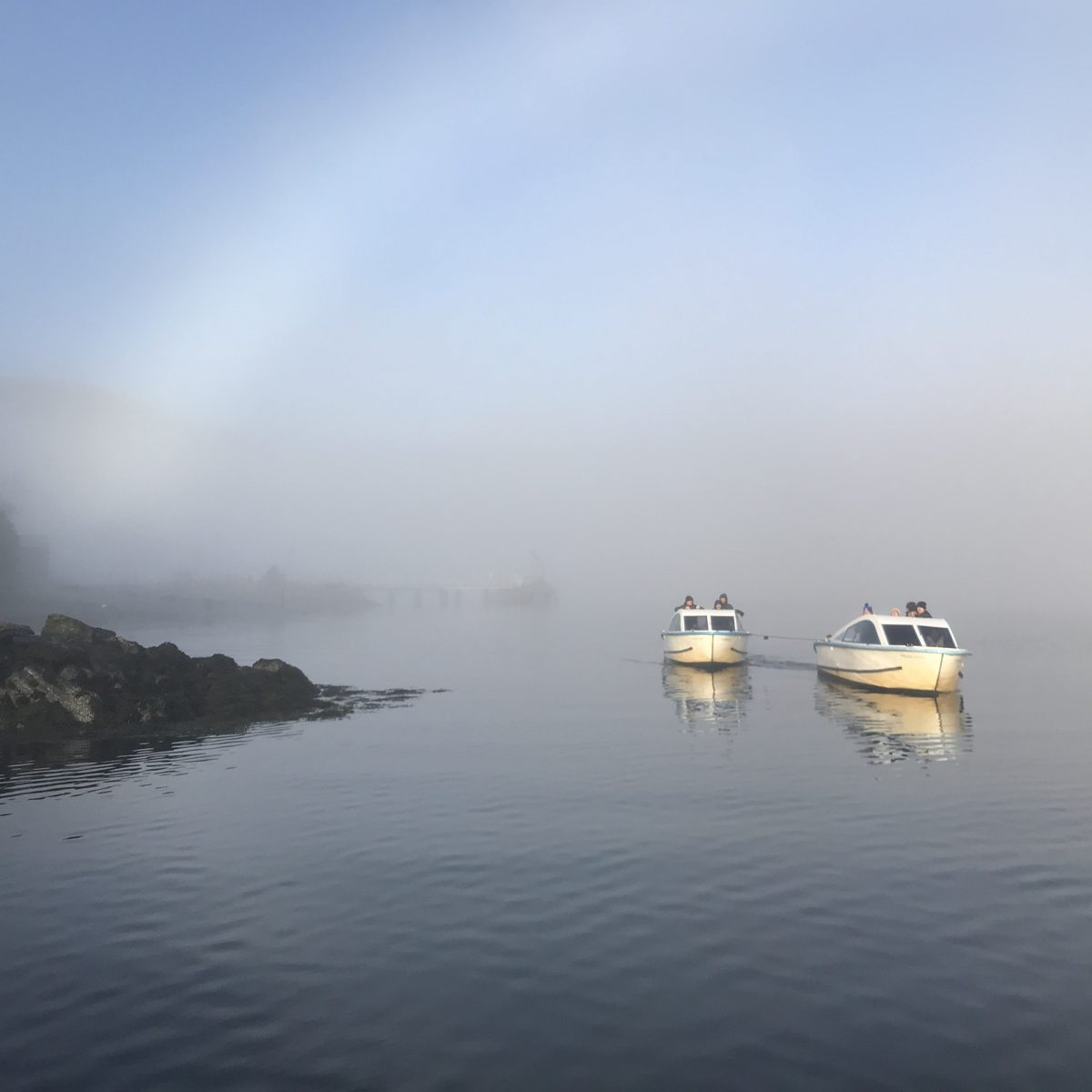 Two boats on calm water in foggy conditions, with a rocky shore.