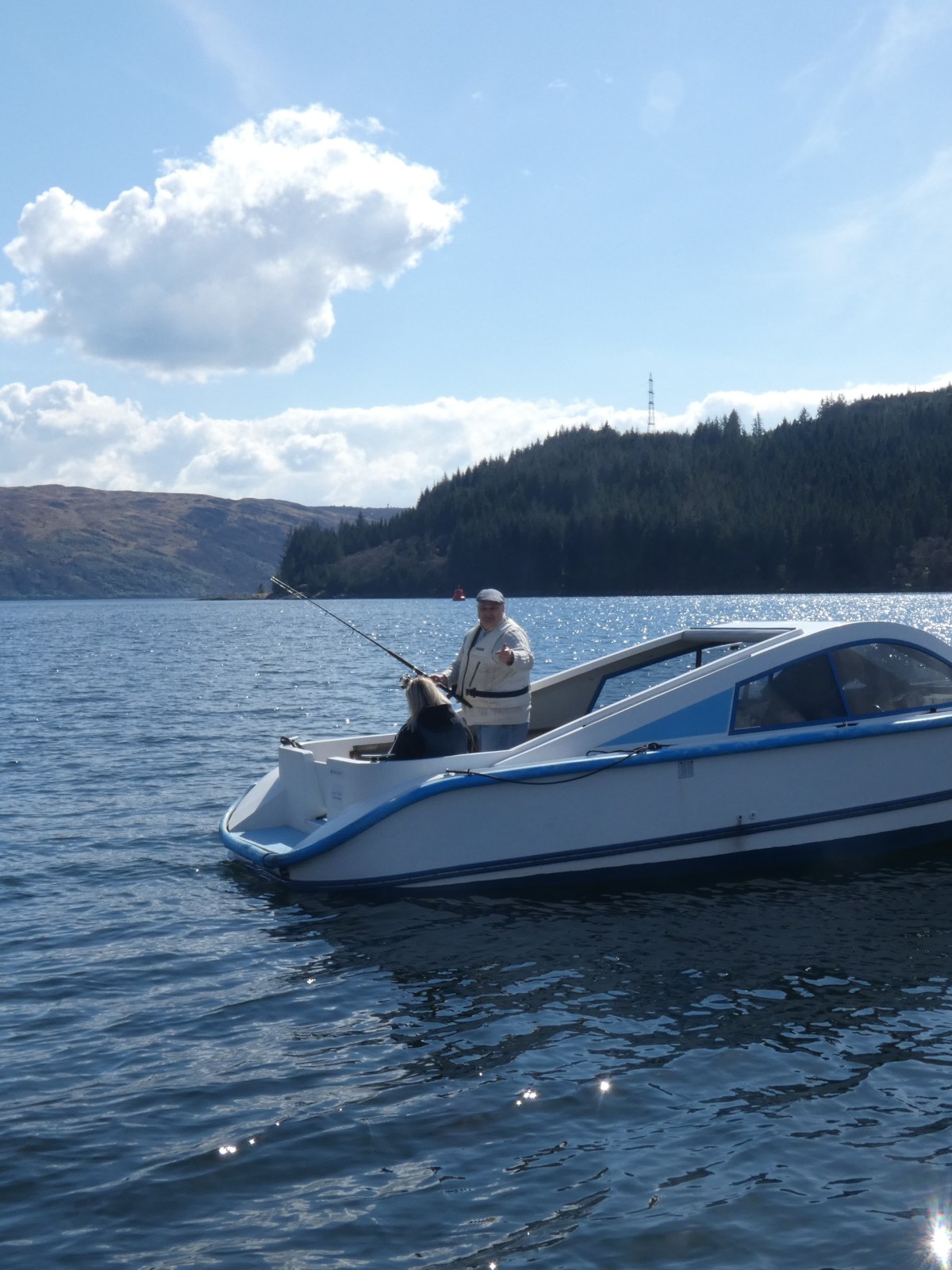 Person in a small white boat fishing on a lake with tree-covered hills in the background.