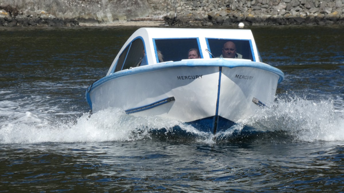 Small motorboat on water with two people, rocky shoreline in background.