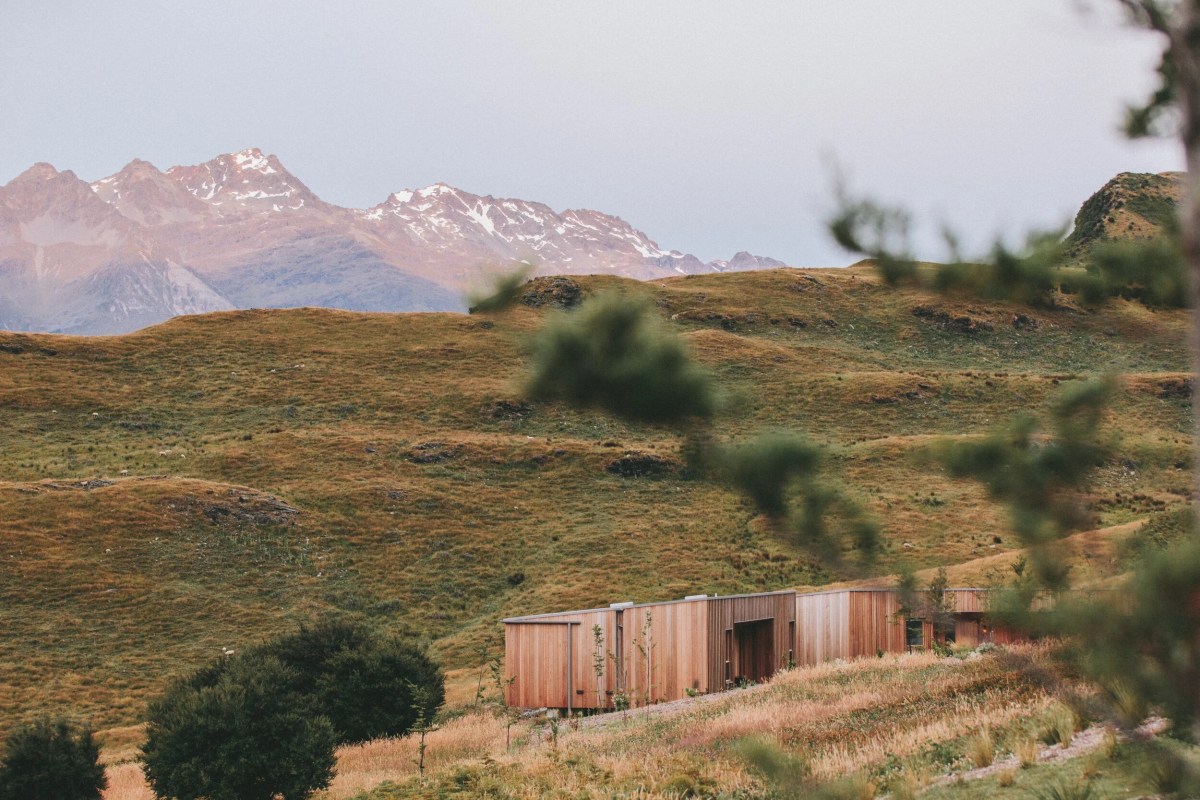 Wooden cabin on grassy hills with distant snow-capped mountains.